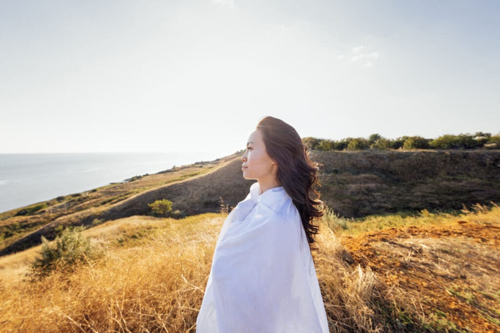 A young handsome Asian woman wrapped in a white sheet and looks at the seascape. A charming Korean woman enjoys nature.
